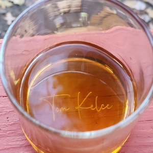 May include: A clear glass with a golden liquid inside. The glass has the name "Tom Kelce" etched into the bottom.