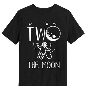 Puede incluir: Camiseta negra con un gráfico blanco. El gráfico presenta la palabra "TWO" sobre una luna con una cara, un astronauta y las palabras "THE MOON". El diseño es simple y juguetón.