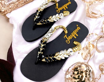 Könnte beinhalten: Ein Paar schwarze Flip-Flops mit goldenen und kristallenen Verzierungen. Die Riemen sind mit klaren und schwarzen Edelsteinen verziert, und die Fußbetten tragen den Schriftzug "Inoppori" in Gold. Die Sandalen liegen auf einer weißen Oberfläche.