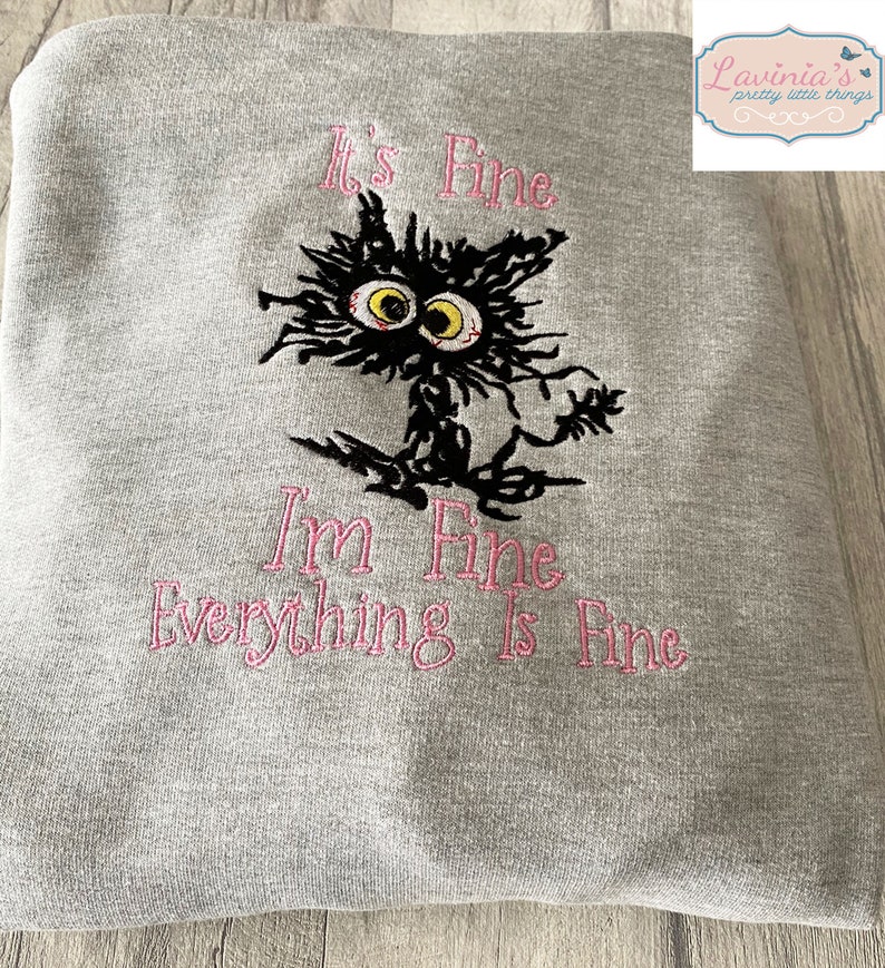 May include: A gray sweatshirt with the embroidered phrase "It's Fine, I'm Fine, Everything Is Fine" in pink. A black cat with yellow eyes and a frazzled appearance is embroidered above the phrase.