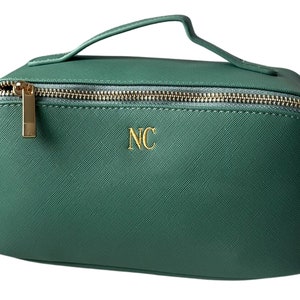 May include: A teal green cosmetic bag with a gold zip and handle. The bag has the gold initials "NC" on the front. The zip and handle hardware are gold. The bag is rectangular with rounded edges.