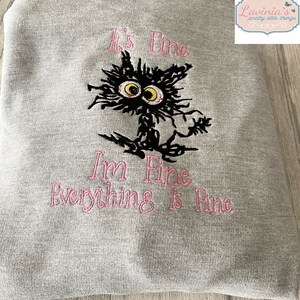 May include: A gray sweatshirt with the embroidered phrase "It's Fine, I'm Fine, Everything Is Fine" in pink. A black cat with yellow eyes and a frazzled appearance is embroidered above the text.