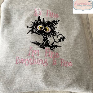May include: A gray sweatshirt with the embroidered phrase "It's Fine, I'm Fine, Everything Is Fine" in pink. A black cat with yellow eyes and a frazzled appearance is embroidered above the text.