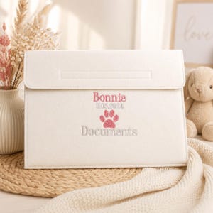 May include: A cream-coloured felt document folder featuring embroidered text that reads Bonnie, 11.05.2024, and Documents, accompanied by a pink paw print graphic. The folder has a rectangular flap closure and a clean, structured design.