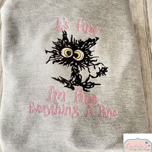 May include: A gray sweatshirt with the embroidered phrase "It's Fine, I'm Fine, Everything Is Fine" in pink. The design features a distressed black cat with yellow eyes and a frazzled appearance.
