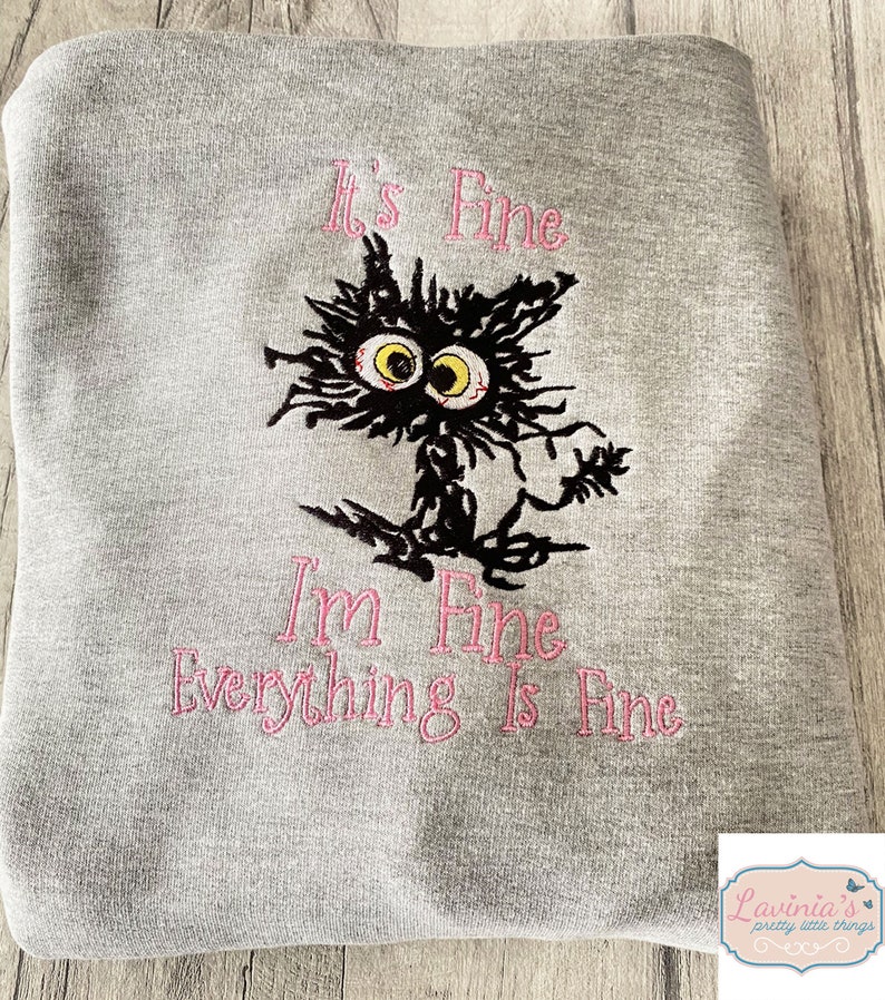 May include: A gray sweatshirt with the embroidered phrase "It's Fine, I'm Fine, Everything Is Fine" in pink. The design features a distressed black cat with yellow eyes and a frazzled appearance.