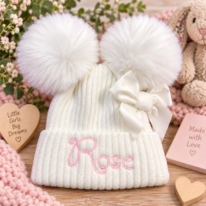 Op de afbeelding: Een witte geribbelde gebreide muts met twee grote witte imitatiebont pompons aan de bovenkant, een witte satijnen strik aan de zijkant en de naam Rose geborduurd in roze cursief schrift op de voorste boord.