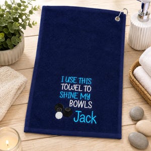 May include: A navy blue rectangular towel featuring an embroidered design with the white and light blue text "I USE THIS TOWEL TO SHINE MY BOWLS Jack" and illustrations of three lawn bowls. The towel includes a silver metal grommet and hook.