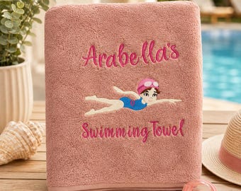 Könnte beinhalten: Ein altrosa Badetuch mit dem eingestickten Text Arabella's Swimming Towel in dunkelrosa Schreibschrift. Zwischen den Textzeilen befindet sich ein gesticktes Motiv einer schwimmenden Person mit blauem Badeanzug und pinker Badekappe.