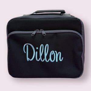 Puede incluir: Una bolsa de almuerzo rectangular negra con un asa superior y un bolsillo frontal con cremallera. El nombre Dillon est&aacute; bordado en letras cursivas de color azul claro en la parte frontal. La bolsa tiene bordes gris oscuro y dos tiradores a juego.