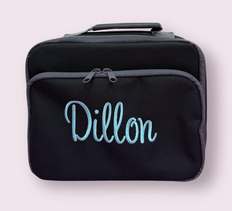 Puede incluir: Una bolsa de almuerzo rectangular negra con un asa superior y un bolsillo frontal con cremallera. El nombre Dillon est&aacute; bordado en letras cursivas de color azul claro en la parte frontal. La bolsa tiene bordes gris oscuro y dos tiradores a juego.