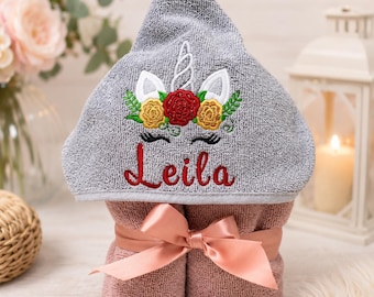 May include: A gray hooded towel featuring an embroidered unicorn design with a floral crown, white ears, and a horn. The name Leila is embroidered in red script below the unicorn. The towel is tied with a dusty pink satin ribbon bow.