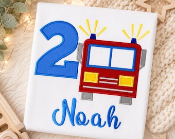 May include: A white shirt featuring an embroidered design of a large blue number two, a red fire truck with yellow headlights and blue lights, and the name Noah in blue cursive script. The design includes yellow stitched light rays above the truck.