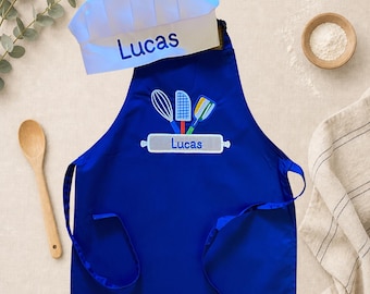 May include: A blue apron and chef's hat set with the name "Lucas" embroidered on both. The apron features embroidered kitchen tools and a rolling pin design. A wooden spoon and small bowl of flour are also visible.