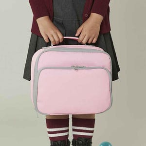 Puede incluir: Una bolsa de almuerzo rectangular en un suave color rosa pastel con cierre de cremallera gris y ribete gris en los bordes. La bolsa incluye un asa superior para transportarla y un bolsillo frontal grande con cremallera, ofreciendo un dise&ntilde;o estructurado.