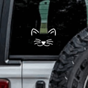 May include: White cat face decal on a vehicle window. The decal features a simple outline of a cat's face, including ears, eyes, nose, and whiskers. The image is set against a dark background, with a tire visible on the right.