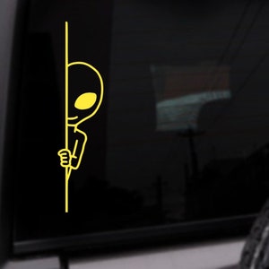 May include: A yellow alien sticker peeking around a corner. The alien has a large head, small body, and a big smile.