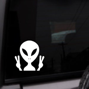 May include: White alien sticker with large oval eyes and a small mouth, giving a peace sign with both hands. The sticker is on a black background, likely a car window.