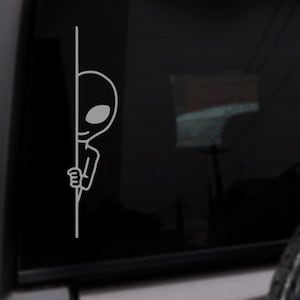 May include: A black and white sticker of an alien peeking around a corner. The alien is drawn in a simple cartoon style.