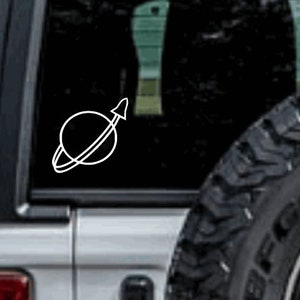 May include: A white vinyl sticker of a planet with a rocket ship orbiting it. The sticker is on the back window of a silver car.