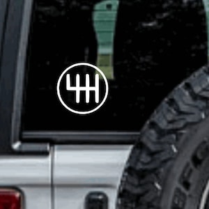 May include: A white vinyl sticker with a black outline of a 4-speed manual transmission gear shift pattern. The sticker is on the back window of a silver car.