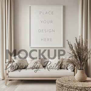 May include: A white frame mockup with the text "PLACE YOUR DESIGN HERE" on a beige wall. The frame is above a light beige couch with three pillows. A wicker side table and a vase with dried branches are in front of the couch.