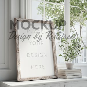 May include: A white framed mockup poster with the text "Place your design here" on a white dresser with a potted plant and books.