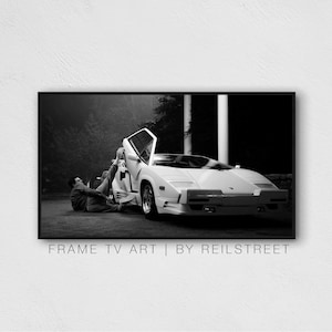 May include: A black and white photograph of a person sitting next to a white Lamborghini Countach with the door open.