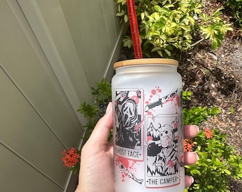 May include: White frosted tumbler with a bamboo lid and a red straw. The tumbler features a graphic of four horror movie characters: Ghost Face, Jason Voorhees, Michael Myers, and Freddy Krueger. The text "Ghost Face", "The Camper", and "The Shape" are printed on the tumbler.