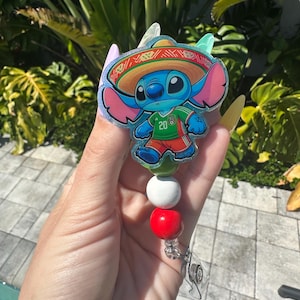 May include: A badge reel featuring a blue cartoon character wearing a sombrero and a green and red jersey. The badge reel has a white, red, and green beaded accent. The character is holding a soccer ball.
