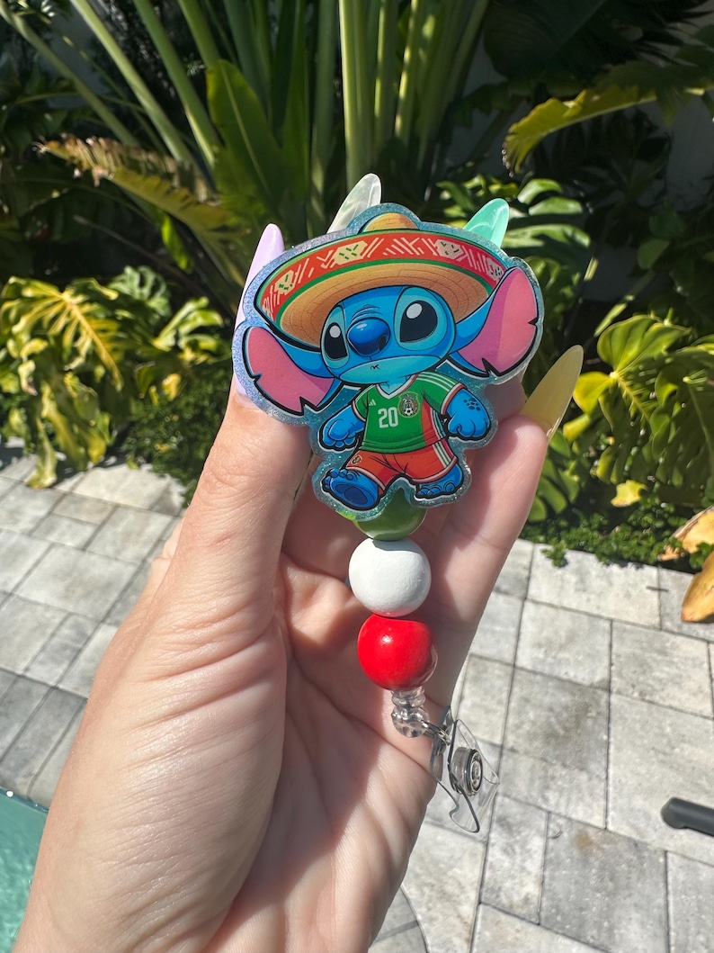 May include: A badge reel featuring a blue cartoon character wearing a sombrero and a green and red jersey. The badge reel has a white, red, and green beaded accent. The character is holding a soccer ball.
