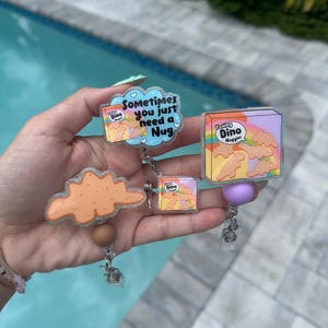 May include: A collection of badge reels featuring a cartoon box of "Yummy Dino Nuggets" and a cloud-shaped nugget. One reel has the text "Sometimes you just need a Nug". The reels are clear with glitter accents and a purple or brown bead.