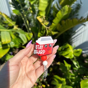 May include: Red and white cartoon-style coffee cup with the text "Stay Sharp" and a smiley face. The cup is attached to a retractable badge holder with red and white beads.