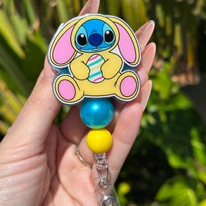 May include: A blue Stitch character wearing a yellow bunny costume and holding a striped Easter egg. The character is attached to a blue and yellow beaded retractable badge holder.