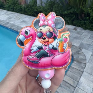May include: A badge reel featuring a glittery acrylic topper of a cartoon mouse wearing heart-shaped sunglasses and a pink polka-dot bow, sitting in a pink flamingo pool float while holding a glass of orange juice with a strawberry garnish.