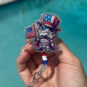 May include: A badge reel featuring a cartoon Stormtrooper character wearing a patriotic Uncle Sam hat and holding a small American flag. The reel has a red, white, and blue beaded accent and a clear plastic clip.