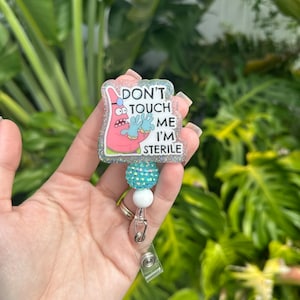 May include: A retractable badge reel with a cartoon image of Patrick Star from Spongebob Squarepants. The badge reel has the text "DON'T TOUCH ME I'M STERILE". The reel has a glittery border, a turquoise beaded accent, and a white bead.