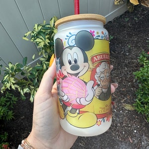 May include: A white tumbler with a bamboo lid and a red straw. The tumbler features a cartoon image of Mickey Mouse holding a pink pastry, with the text "Abuelita Cual Dulce" printed on the side.