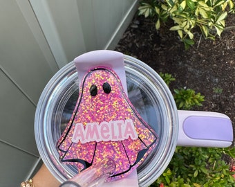 May include: A pink glitter ghost with black eyes and the name "Amelia" written on it. The ghost is attached to a clear lid with a purple handle.