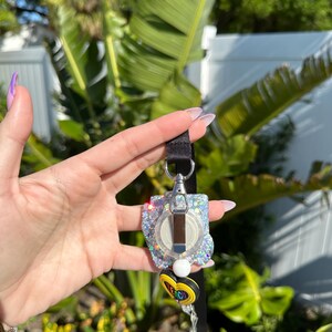 May include: A glittery badge reel with a silver clip and a black lanyard. The reel has a clear, round center with a white and black design, surrounded by iridescent glitter. A charm with the word "TEACH" is attached.