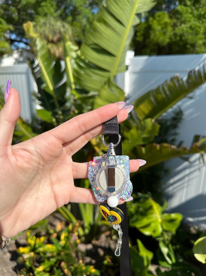 May include: A glittery badge reel with a silver clip and a black lanyard. The reel has a clear, round center with a white and black design, surrounded by iridescent glitter. A charm with the word "TEACH" is attached.
