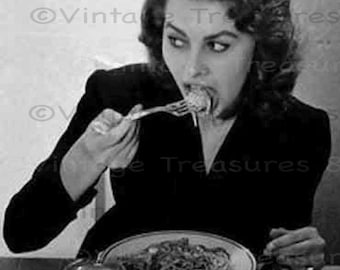 以下が含まれることがあります： 黒いドレスを着た女性がフォークでスパゲッティを食べている白黒写真。彼女はスパゲッティの皿とコップの水のあるテーブルに座っている。