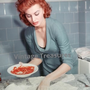 Puede incluir: Una mujer con el pelo rojo está haciendo pizza. Está extendiendo salsa de tomate sobre una masa de pizza. La masa de pizza está sobre una encimera blanca. Hay un plato con salsa de tomate en el fondo.