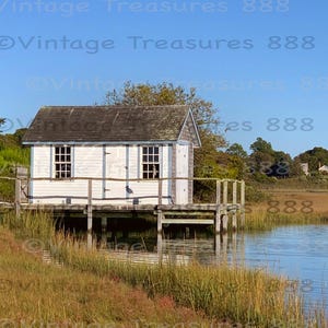 May include: A small, white wooden building with a gray roof sits on a wooden dock over calm water. The building has two windows and a door. The scene is set against a blue sky and marshy landscape.