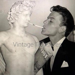 May include: A black and white photo of a man in a suit smoking a cigarette with a marble statue of a man. The man is holding a newspaper with the word "Post" visible.