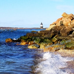May include: A white lighthouse with a black roof stands on a rocky shore. The lighthouse is in the distance, with a blue sky and calm water in the foreground. The water is lapping at the shore, creating small waves.