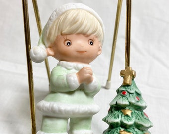 May include: A ceramic figurine of a child in a green outfit and a white hat, with hands clasped in prayer. A small Christmas tree with gold stars and a cross sits beside the child. The figurine is on a white base.