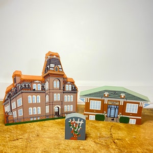 May include: Three wooden miniature buildings, a red brick schoolhouse, a green-roofed library with the text "Cannon Public Library", and a small gray trellis with red flowers.