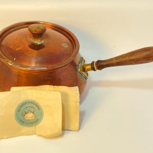 May include: A copper saucepan with a lid and a long wooden handle. A small, circular, light blue paper label is placed in front of the pan, featuring a drawing of a kettle and the text "OLD DUTCH COPPERWARE" and "MADE IN PORTUGAL".