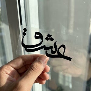 May include: A clear, rectangular sticker with black Arabic calligraphy. The script is stylized and appears to be a word or phrase. The sticker is held up against a window, with natural light illuminating the text.